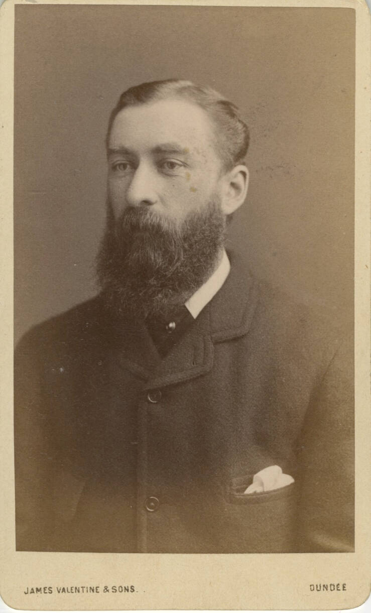 Historic studio portrait photograph of a man from the chest up. He wears a dark, buttoned up jacket and has a beard.